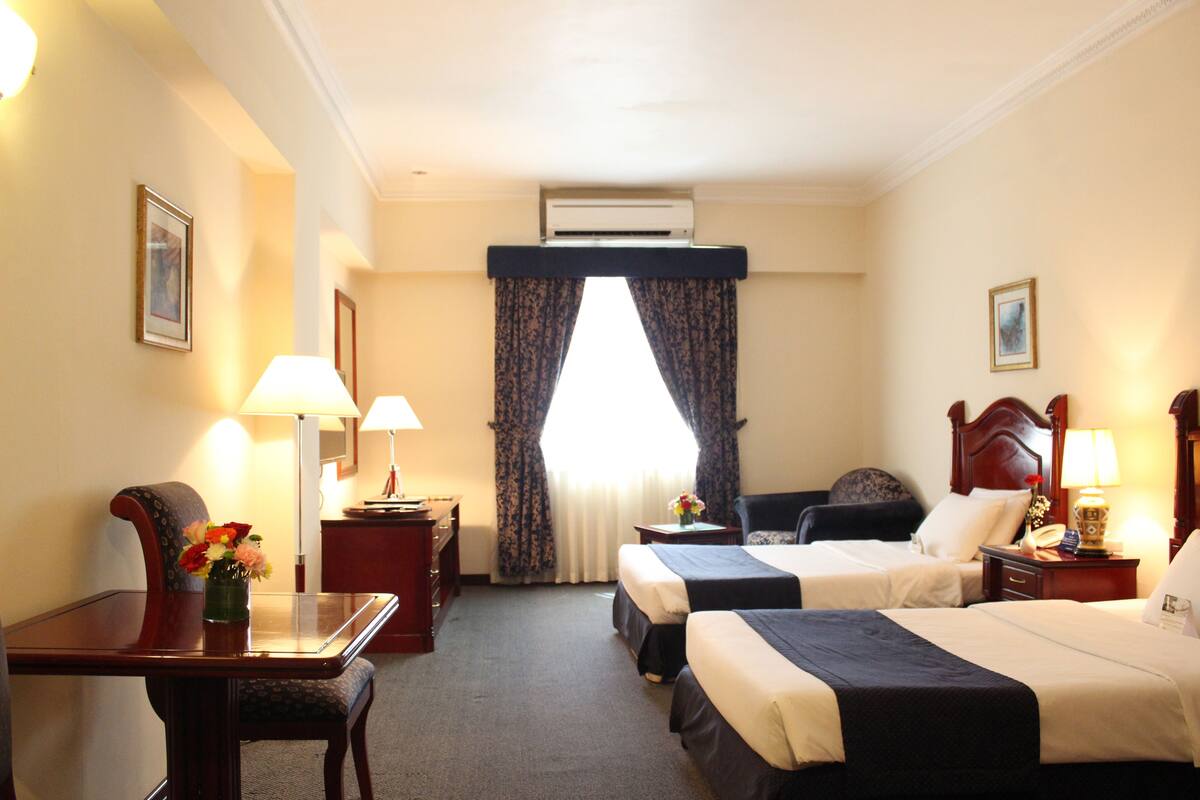 A hotel room with two beds, a desk with a lamp, a chair, a dresser, and an air conditioning unit.