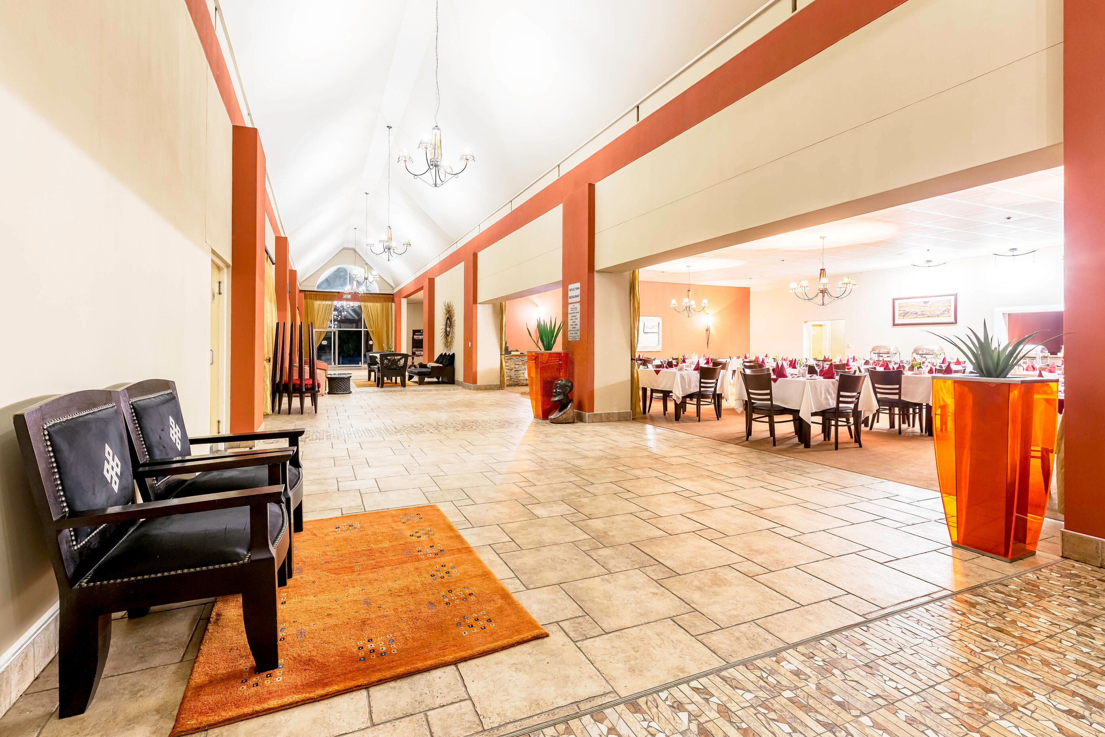 A spacious hotel lobby with a high ceiling, chandeliers, and a central seating area with sofas and a rug. Tables and chairs are arranged for dining in the background.