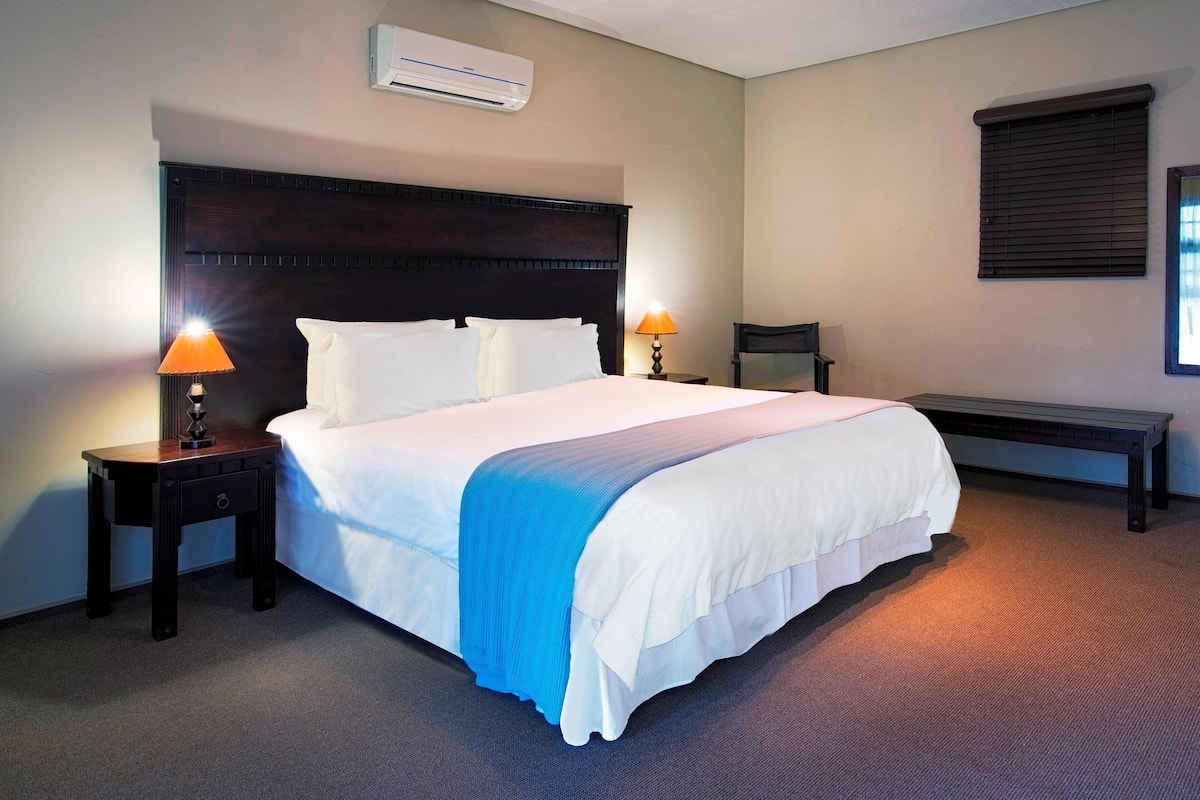 A hotel room with a large bed, two bedside lamps, a bench, and a window with blinds.