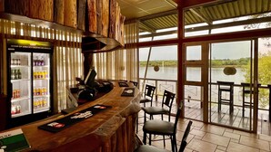 A bar area with a wooden counter, a refrigerator stocked with beverages, and a view of a lake through large windows.