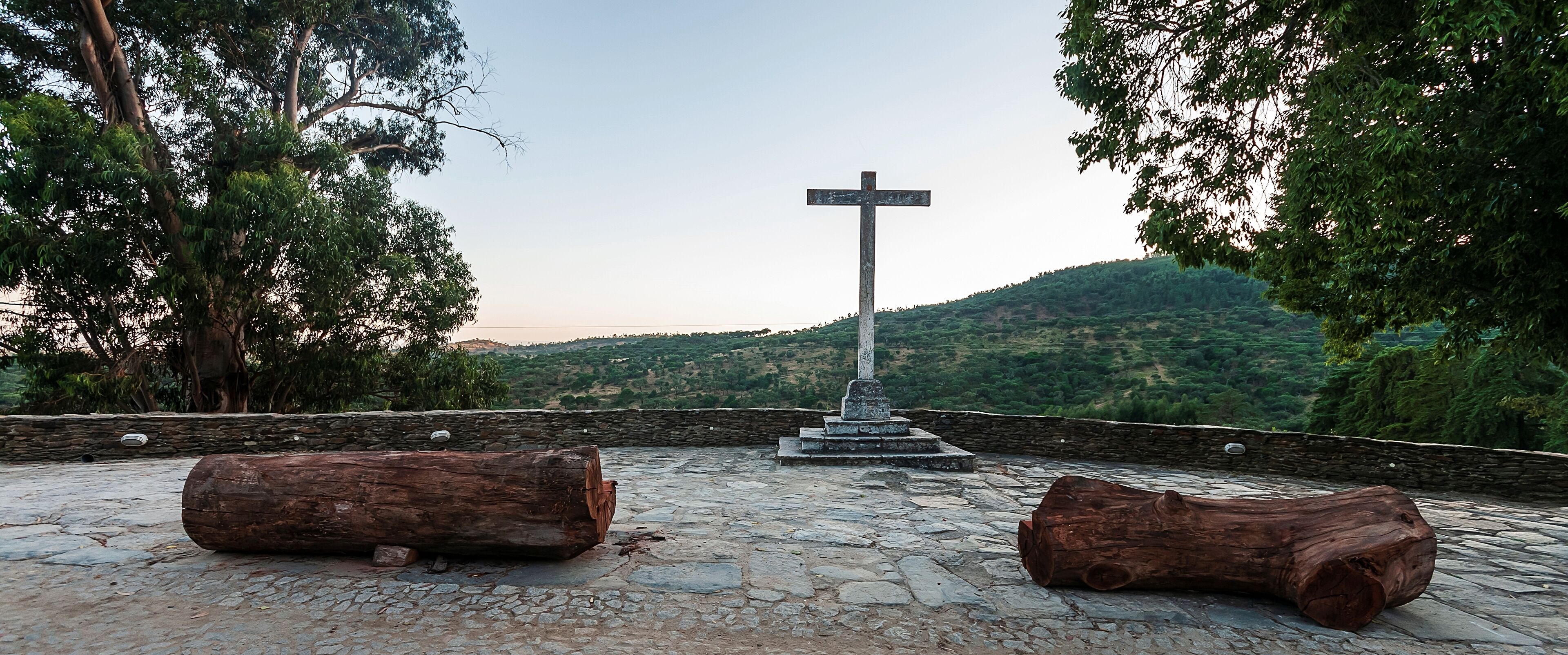 Una cruz de piedra sobre una plataforma de piedra, rodeada de árboles, con vistas a un paisaje montañoso en la distancia.
