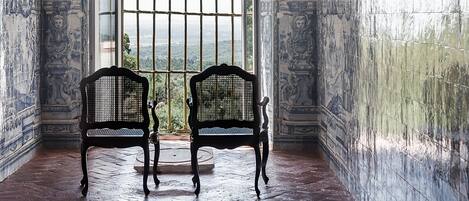 A room with two black wicker chairs facing a large window with metal bars, tiled walls with intricate designs, and a wooden floor.