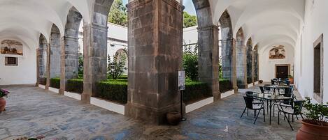 A courtyard with stone flooring, arches, and a central column. Tables and chairs are arranged for outdoor dining.