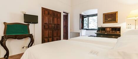 A hotel room with two beds, a television, a wooden wardrobe, a desk with a lamp, and a window with a view.