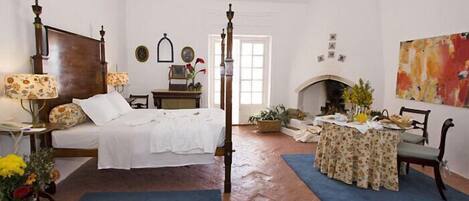 A traditional bedroom with a four-poster bed, a fireplace, a dining table with chairs, and a large abstract painting on the wall.