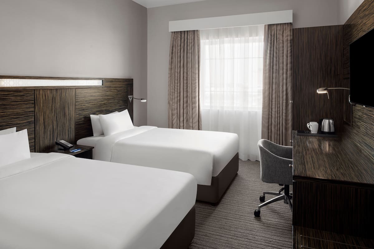 A hotel room with two beds, a desk with a chair, a TV, and a window with curtains.