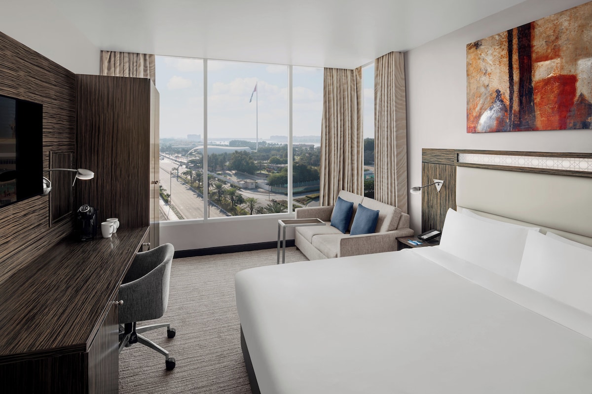 Quarto de hotel com uma cama grande, escrivaninha, cadeira, televisão e vista para a paisagem urbana.