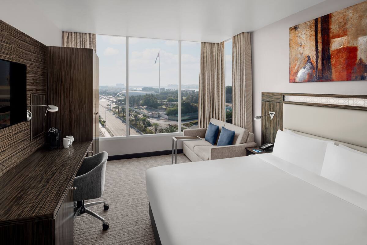 A hotel room with a large bed, a desk, a chair, a sofa, and a view of a cityscape through large windows.