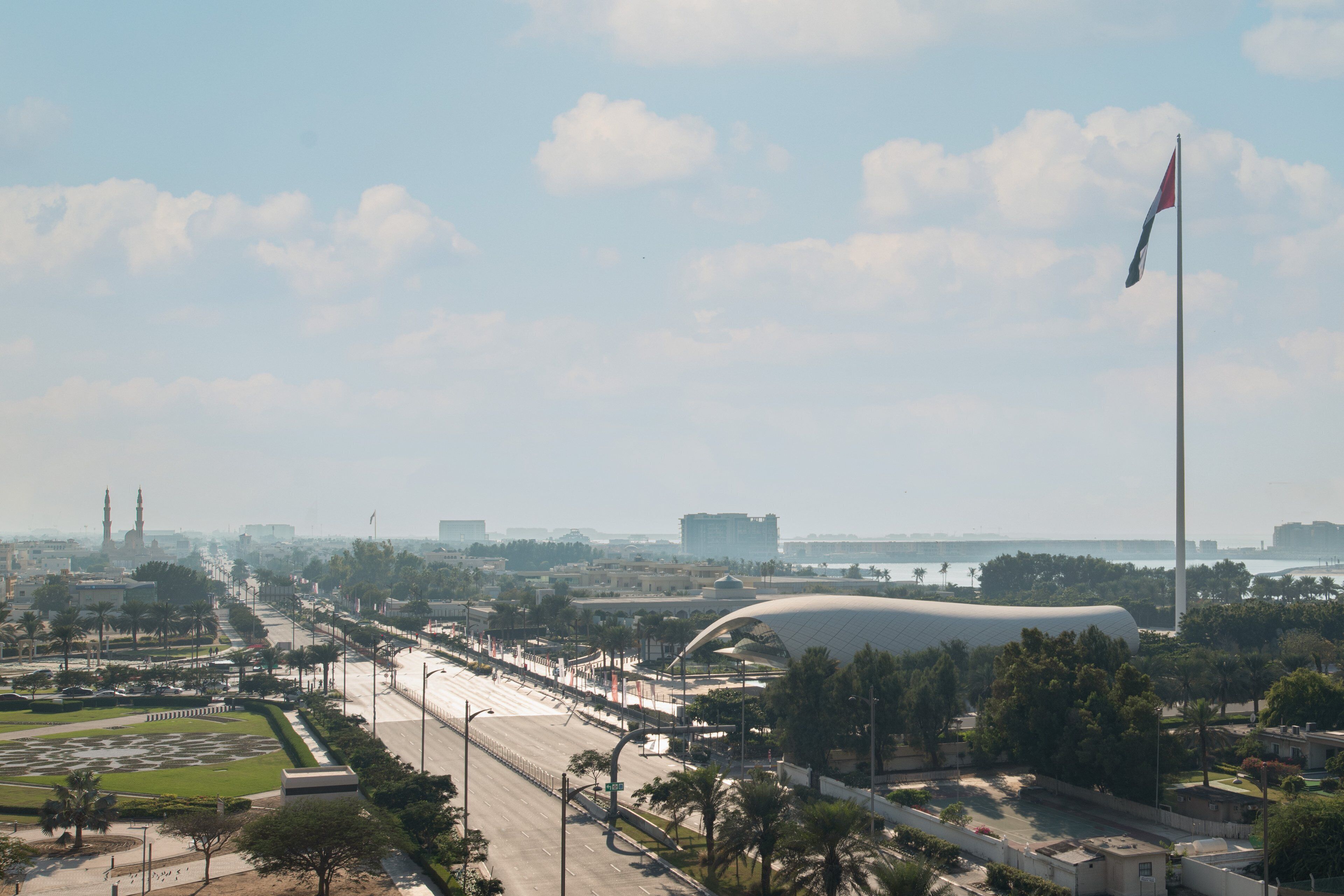 A wide city street with a modern building, a flag, and a mosque in the distance.