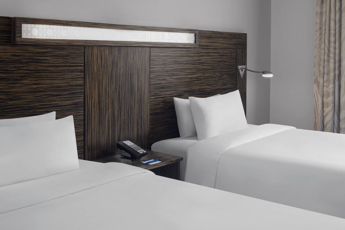 A hotel room with two beds, a nightstand with a telephone, and a wall-mounted light fixture.
