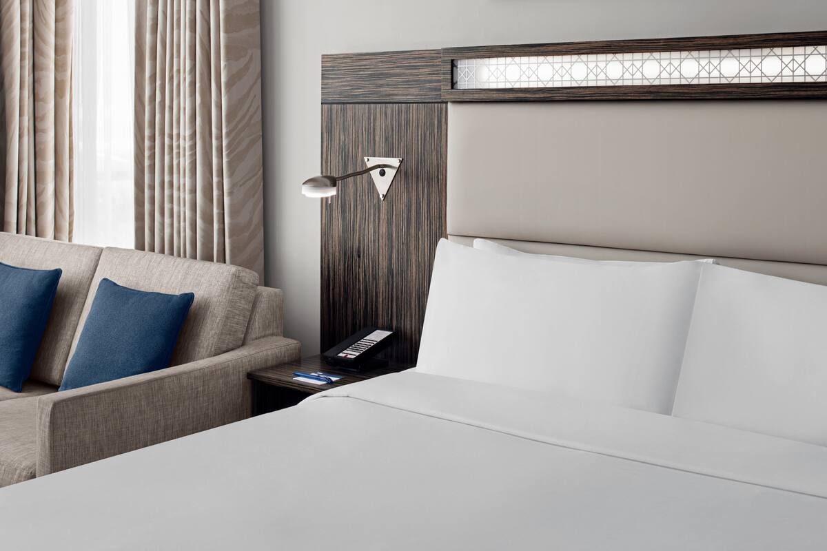 A hotel room with a bed, a nightstand, a lamp, a sofa with cushions, and a window with curtains.