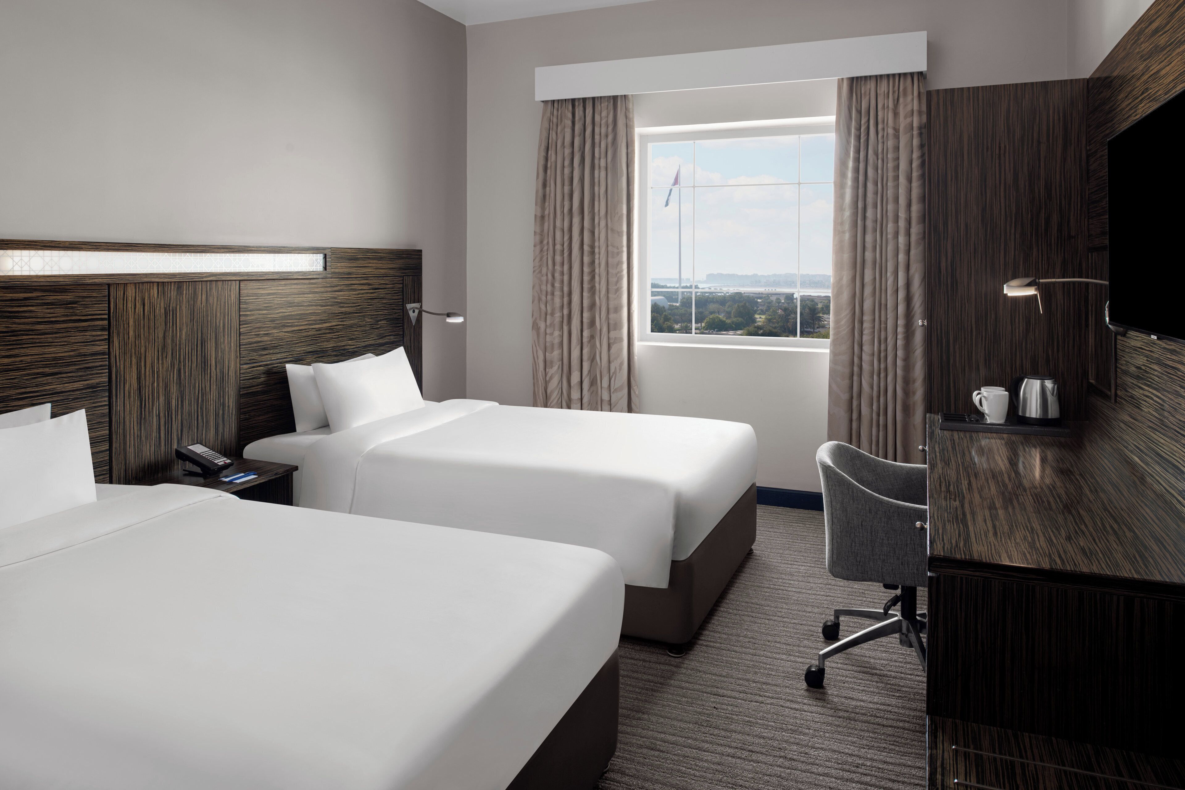 A hotel room with two beds, a desk, a chair, a TV, and a window with curtains.