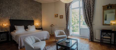 A bedroom with a large bed, two chairs, a glass coffee table, a chandelier, a window with curtains, and a mirror.