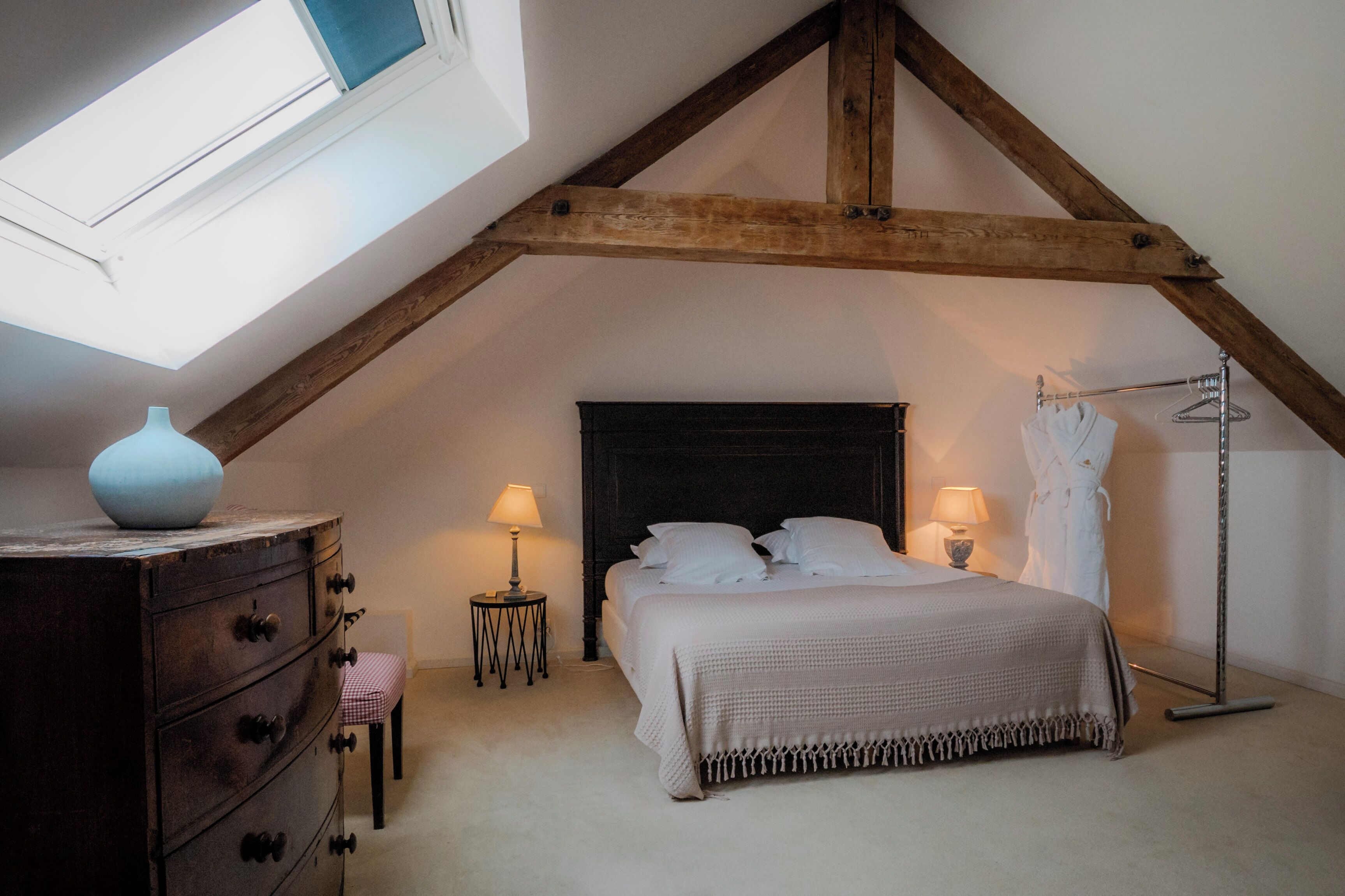 Une chambre avec un plafond en pente, un lit avec une literie blanche, une commode, deux lampes de chevet, un peignoir suspendu à un porte-manteau et un vase posé sur la commode.