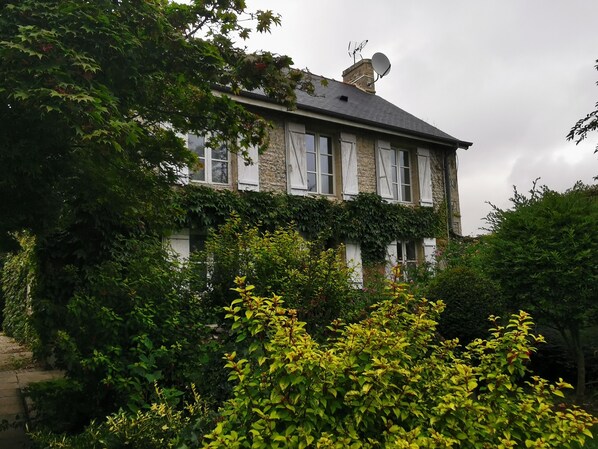 A two-story stone house with white shutters, surrounded by lush greenery and flowering plants.