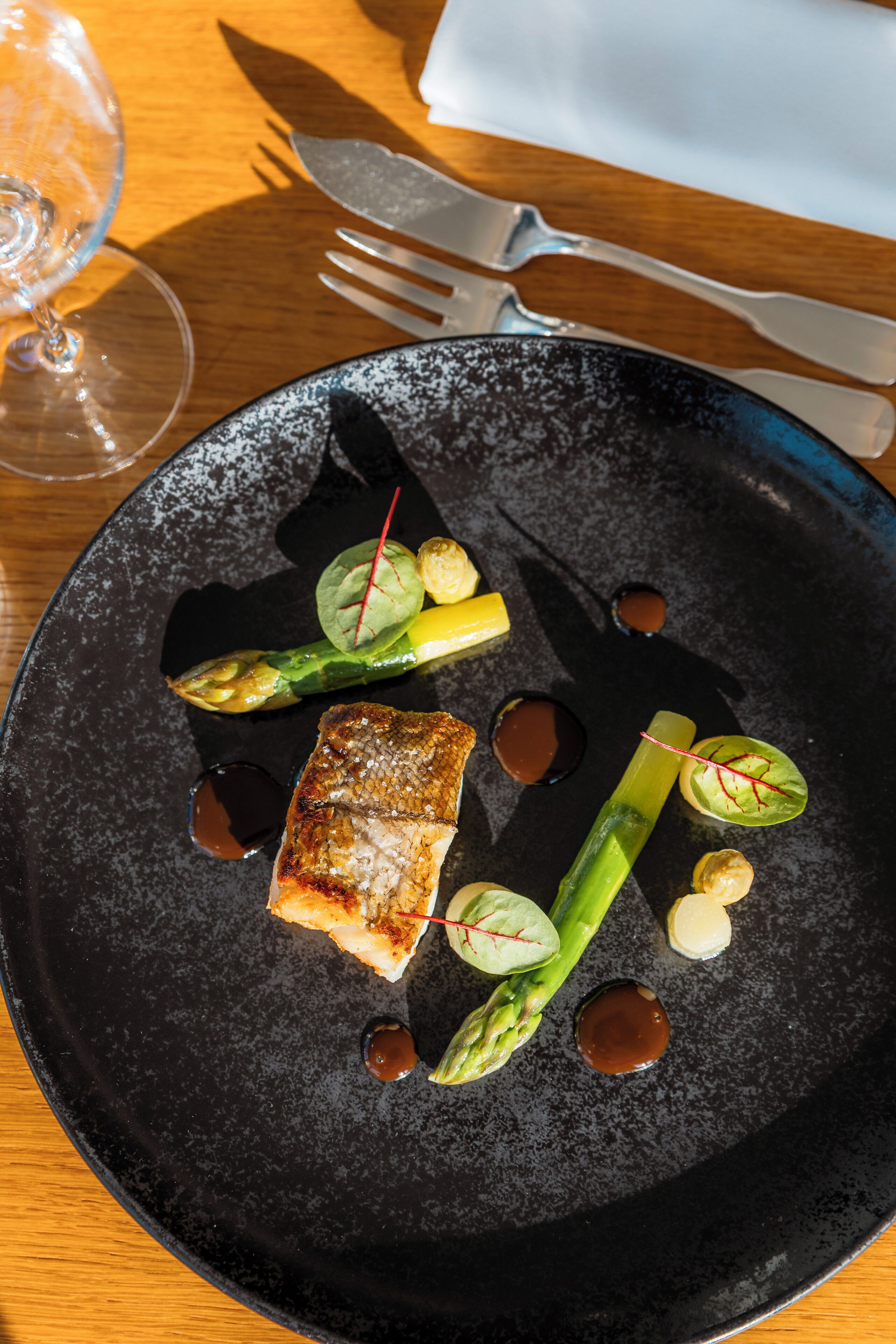 Une assiette contenant un morceau de poisson, des asperges et de petits éléments ronds, possiblement des noix, le tout présenté sur une assiette foncée.