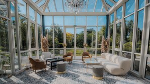 A spacious conservatory with a glass ceiling, a white sofa, two armchairs, a coffee table, and a patterned rug.