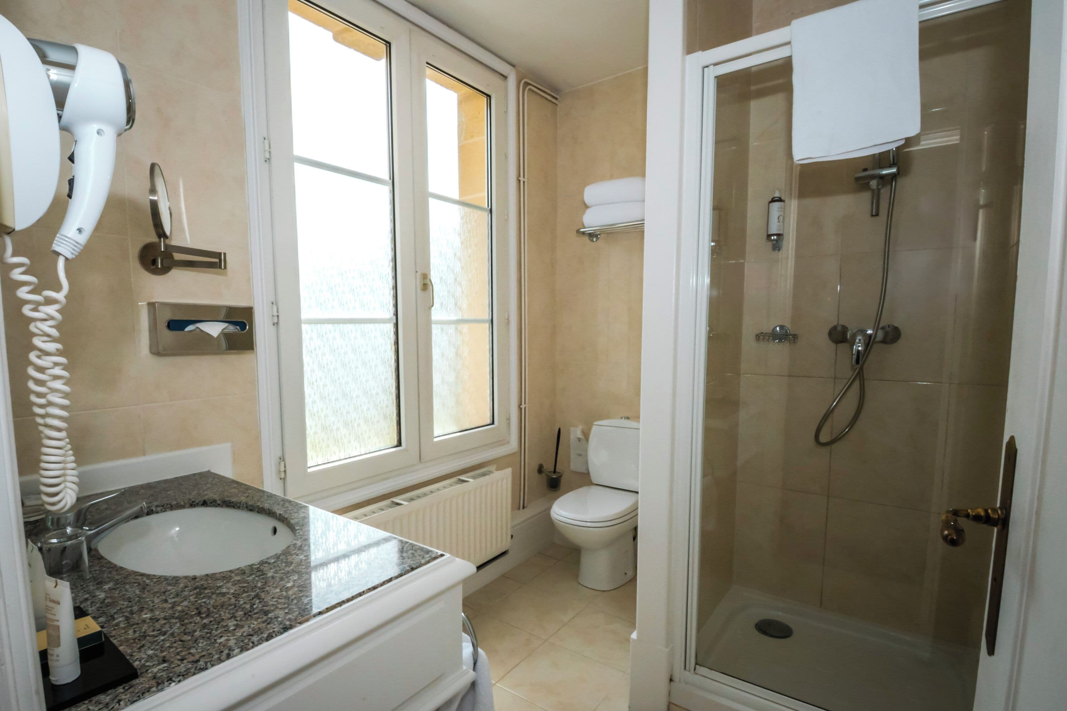 Une salle de bain dotée d’un lavabo avec un plan de toilette en granit, de toilettes, d’une douche à parois de verre et d’un sèche-cheveux fixé au mur.