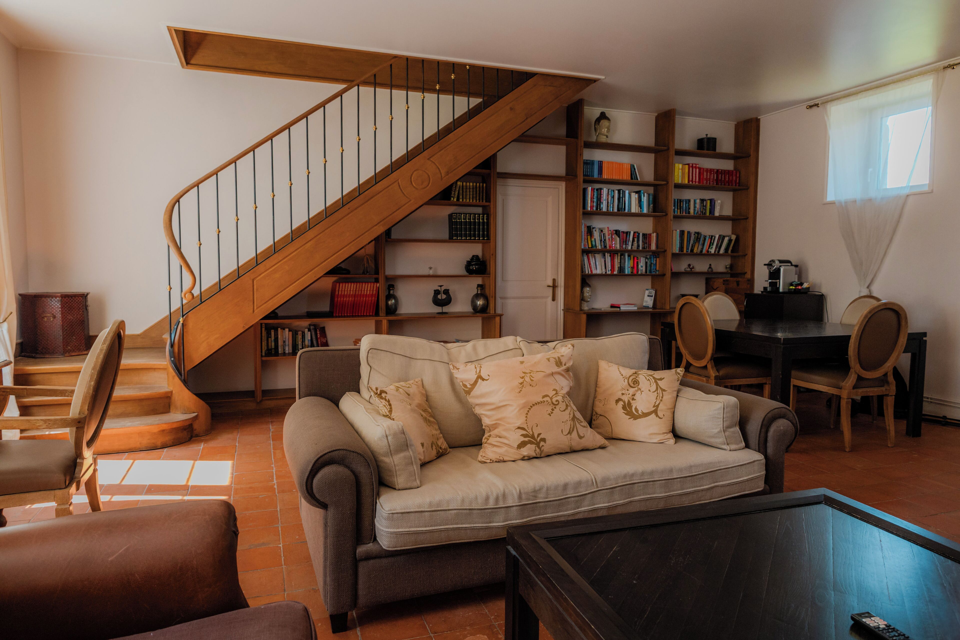 Ein Wohnzimmer mit einer Couch, einem Couchtisch und einer Treppe. Eine Bücherwand voller Bücher und ein Essbereich mit Stühlen sind ebenfalls vorhanden.