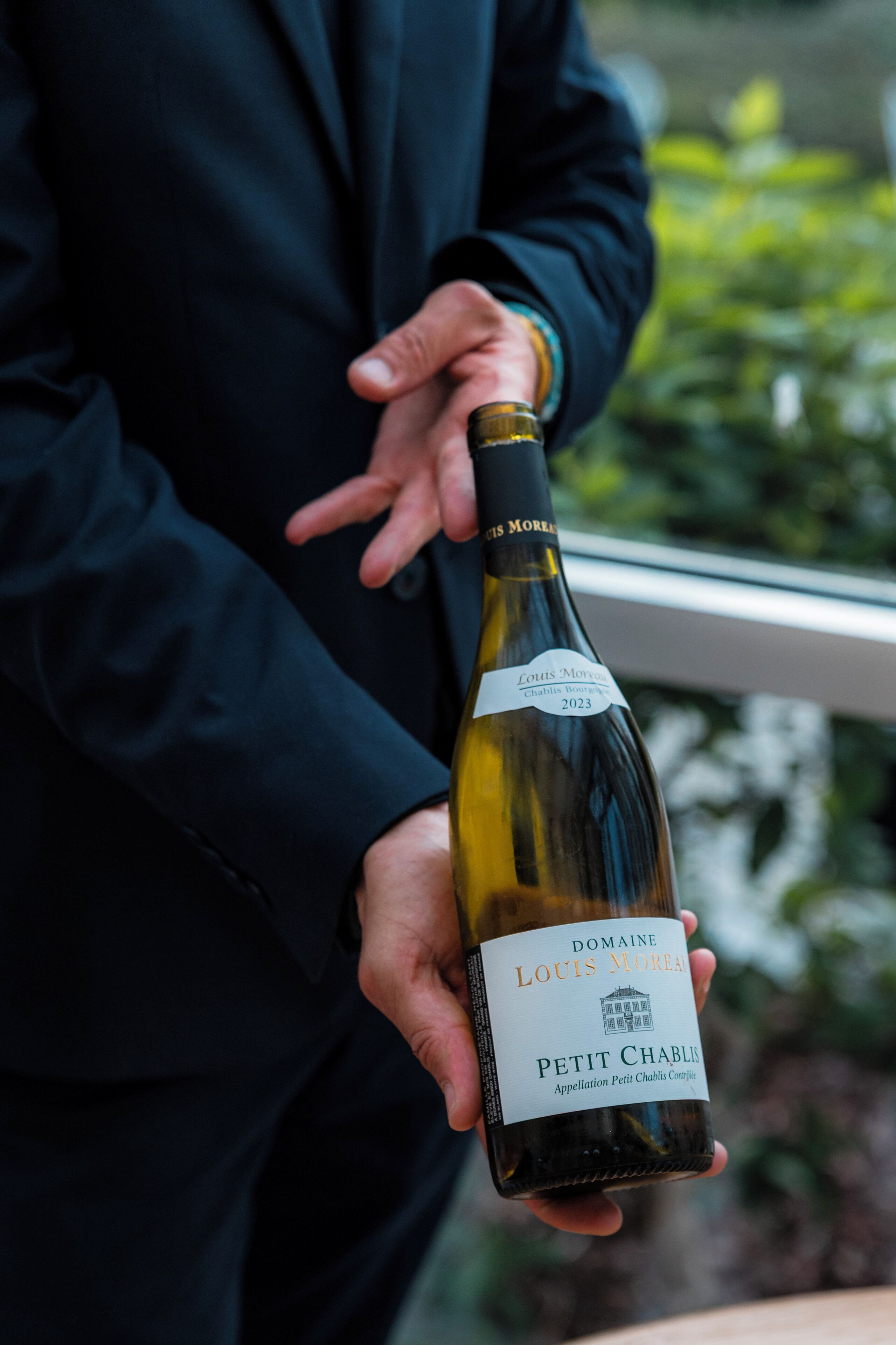 A person holding a bottle of wine with a label that reads "Domaine Louis Moreau Petit Chablis 2023".