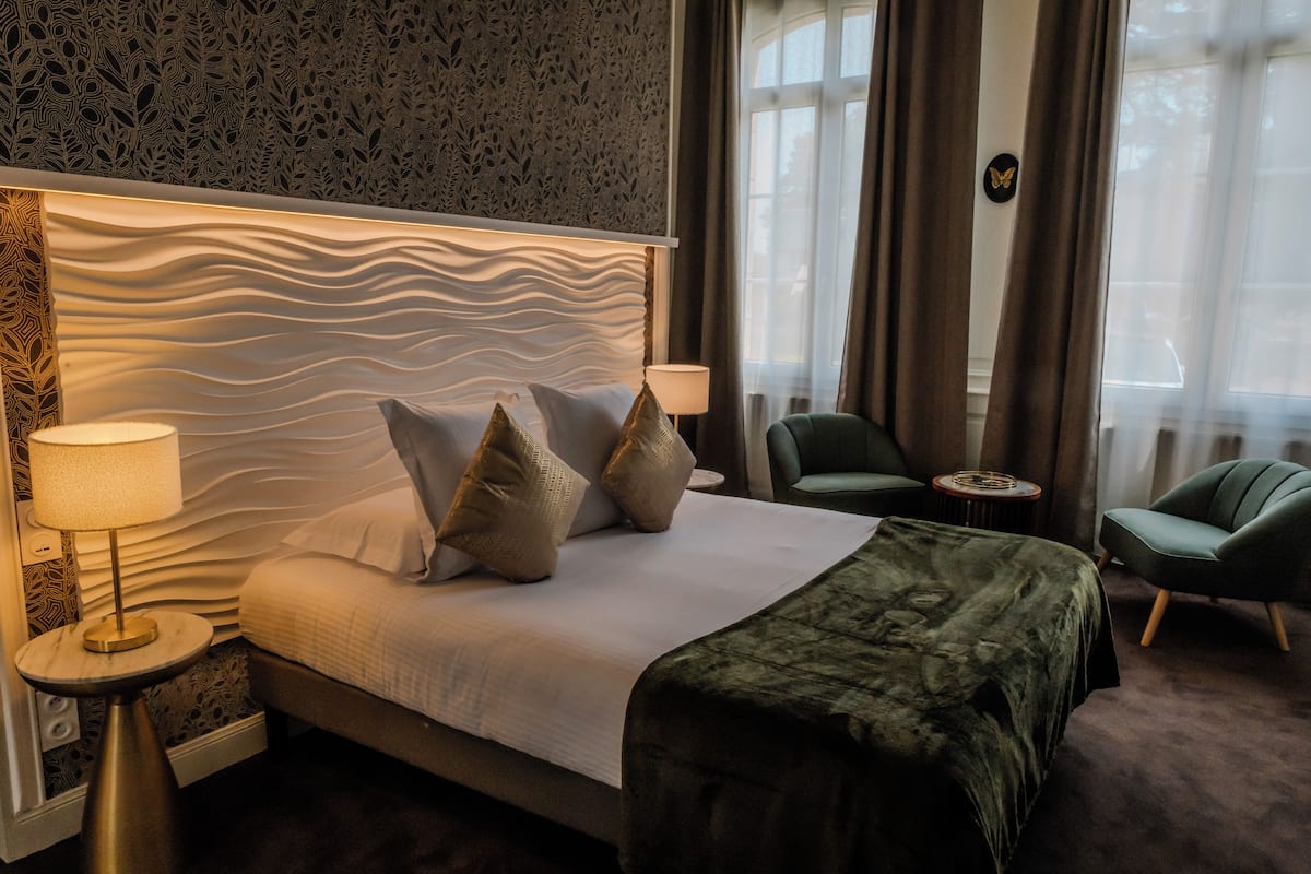Hotellihuone, jossa on moderni sänky, kaksi yöpöytää valaisimineen, vihreä nojatuoli ja suuret ikkunat, joista on näkymä ulos.