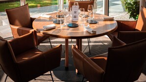 A round dining table with six brown chairs, large windows with a view of a garden, and a stone wall in the background.
