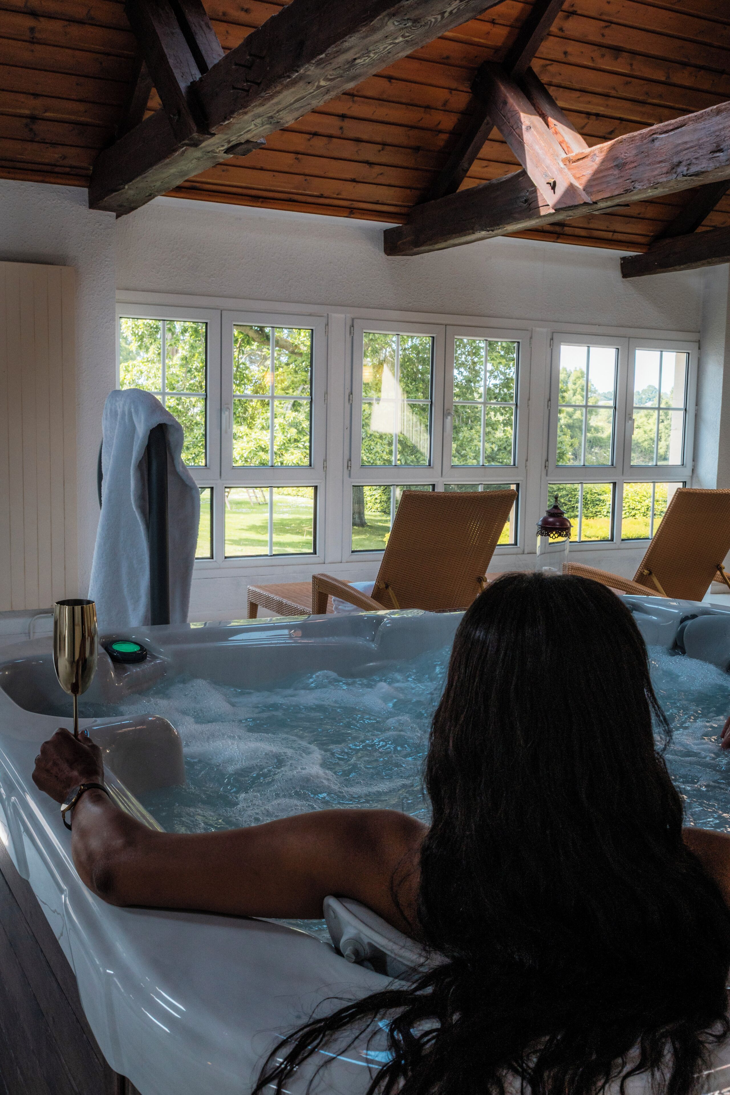 A person relaxing in a hot tub with a drink, surrounded by wicker chairs and large windows.