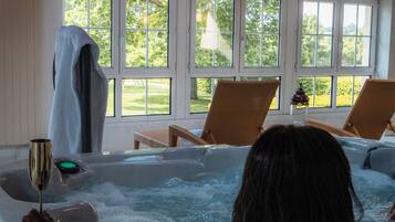 A person relaxing in a hot tub with a drink, surrounded by wicker chairs and large windows.