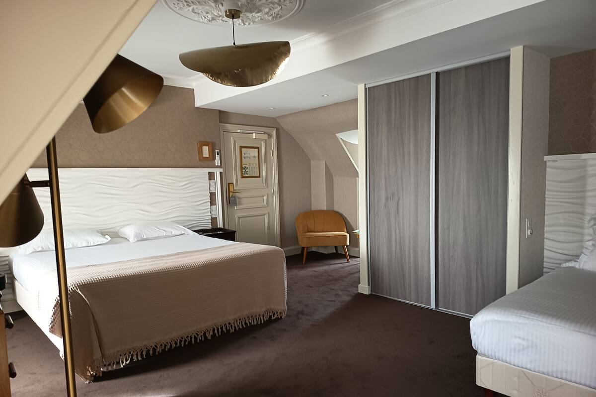 Moderni hotellihuone, jossa on suuri sänky, pienempi sänky, vaatekaappi, tuoli ja kattovalaisin.