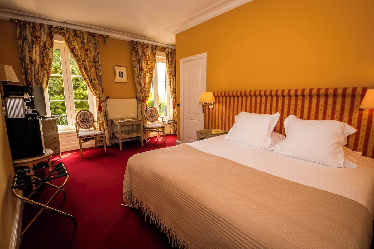 Hotellihuone, jossa on suuri sänky, tuoli, työpöytä lampun kanssa ja näkymä vehreään maisemaan.