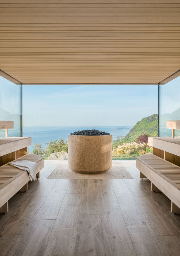 A modern room with a large window, a circular stone fireplace, wooden benches, and a view of the sea. - Lefay Resort & SPA Lago di Garda (Gargnano)