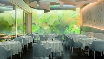 En restaurant med runde borde, grå stole og et stort vindue med et abstrakt grønt motiv.