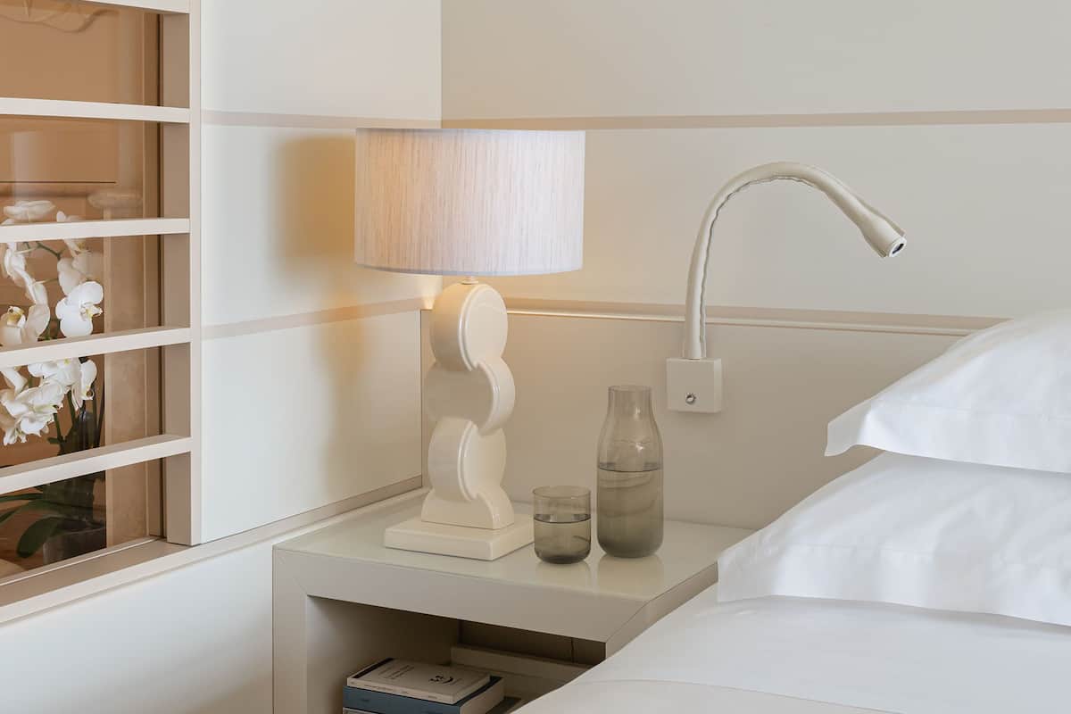 A neatly arranged hotel room with a bed, bedside table, lamp, and a window with blinds.