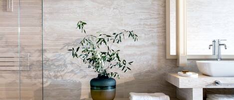 A modern bathroom with a glass shower enclosure, a marble wall, a sink with a mirror, and a vase with green plants.