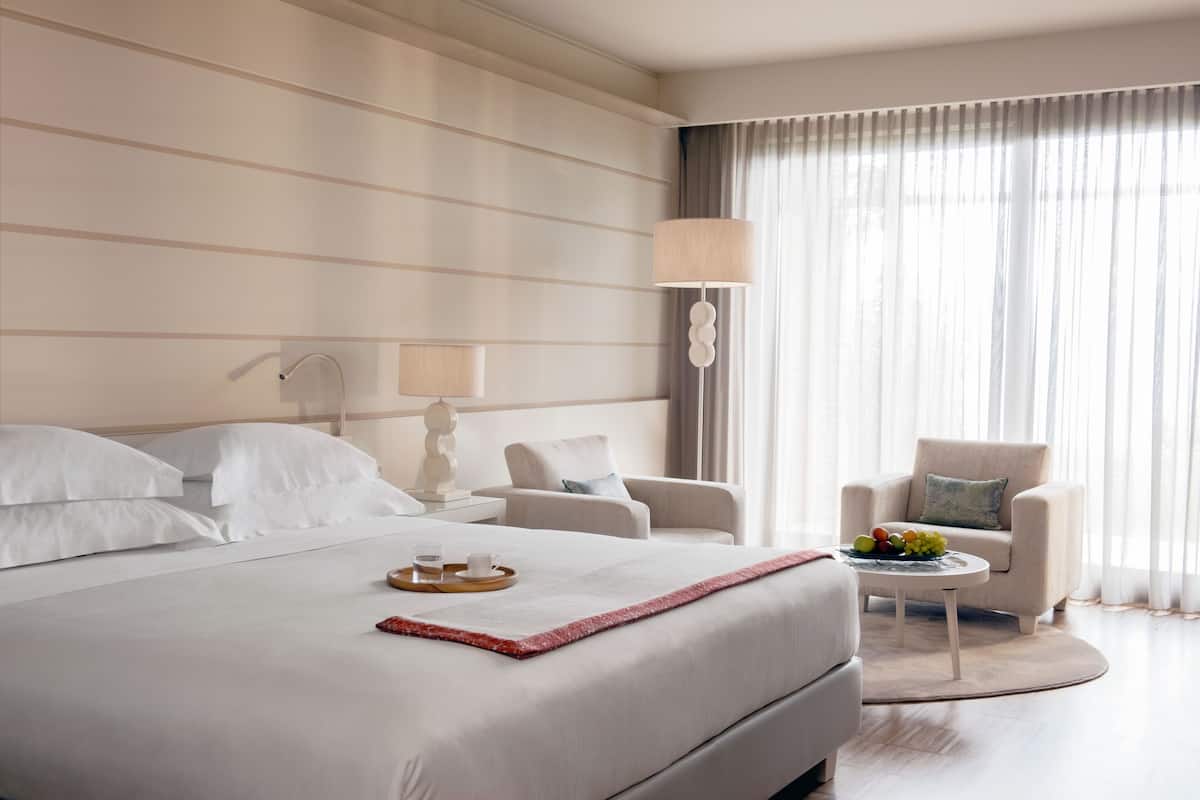 A hotel room with a large bed, two bedside tables, a chair, a small round table with fruit, and a lamp.