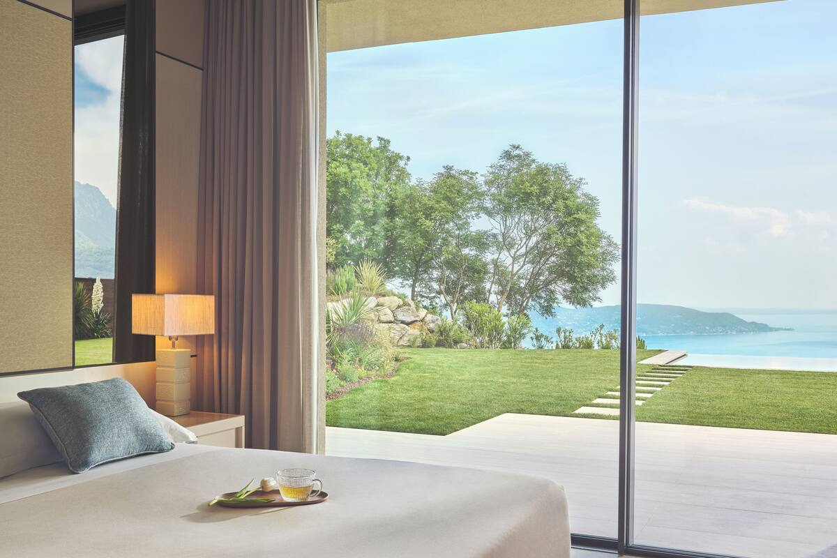 A modern hotel room with a large bed, a bedside table with a lamp, and a view of a lush garden and sea.