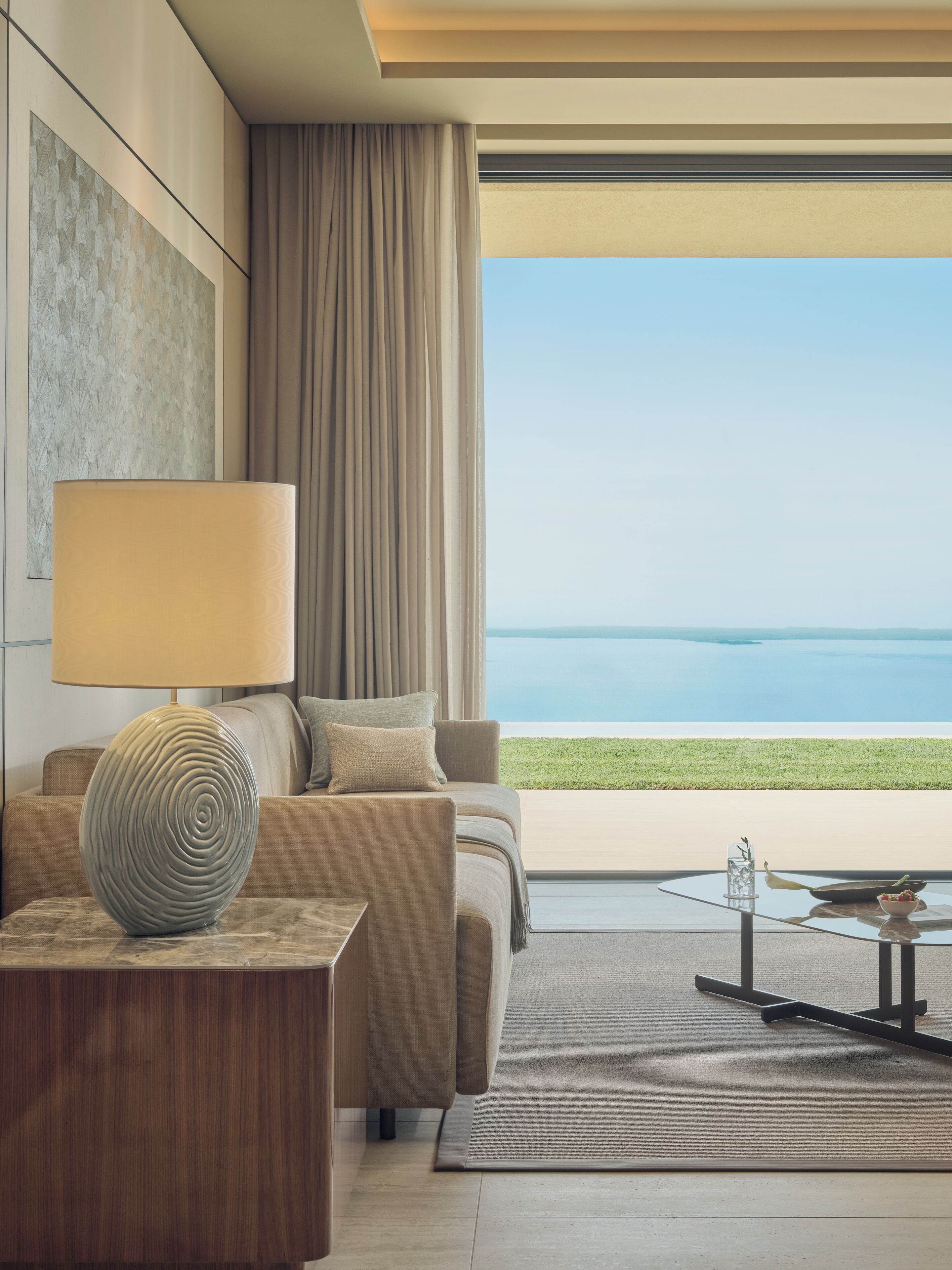 Una sala de estar moderna con un sofá, una lámpara y una mesita de centro. Se ve una ventana grande con vista al mar.