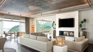 Una sala de estar moderna con un sofá grande, una chimenea y un televisor. Hay ventanas amplias y un balcón con vistas a las montañas.