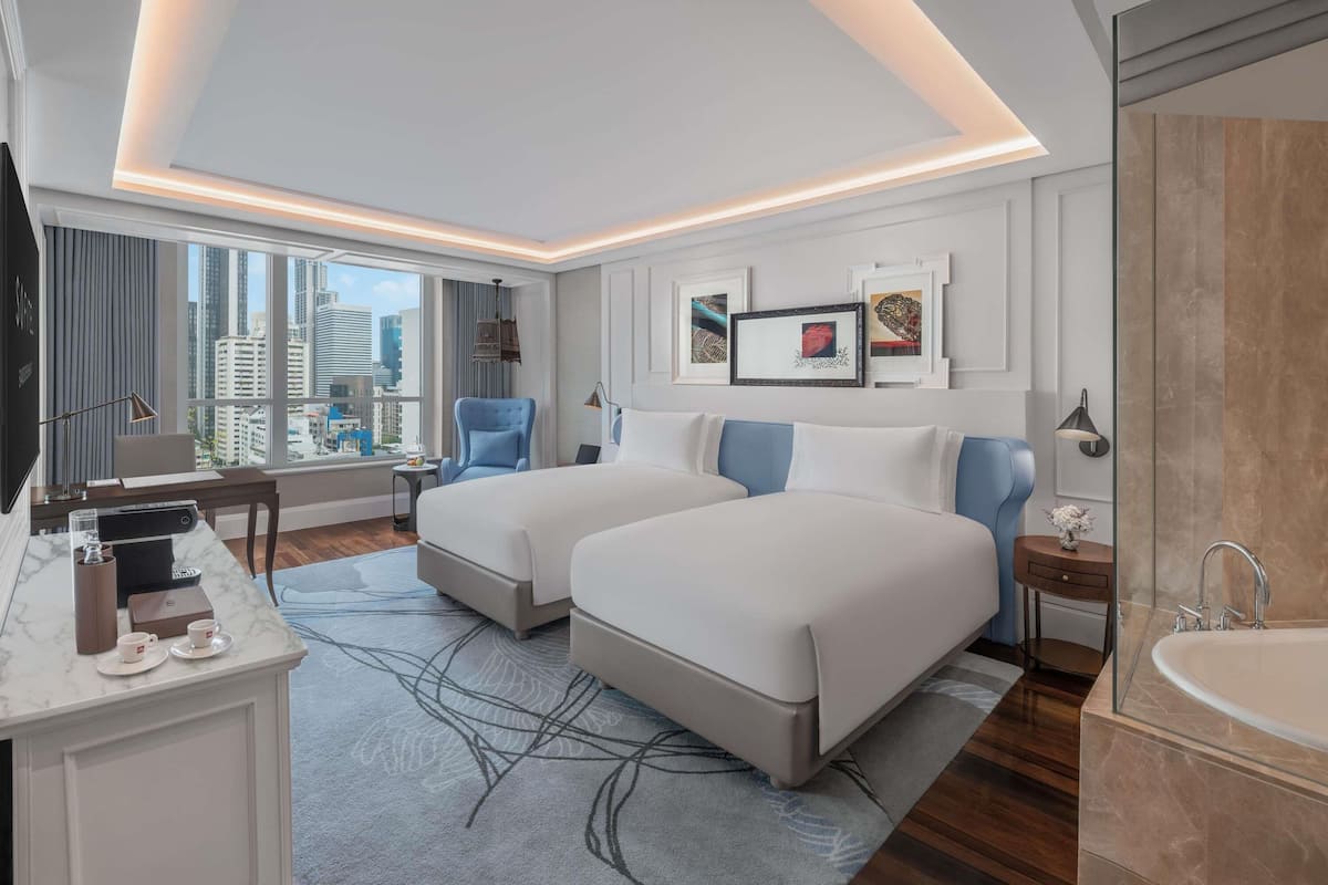 A hotel room with two beds, a desk with a computer, a TV, a large window with a city view, and a bathroom with a bathtub.