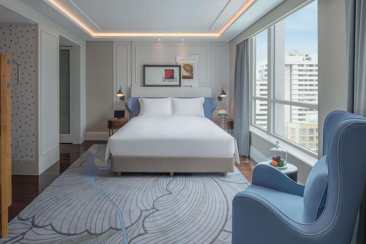A modern hotel room with a large bed, two bedside tables, a nightstand, a blue armchair, a large window with a city view, and a decorative wall with a framed picture.