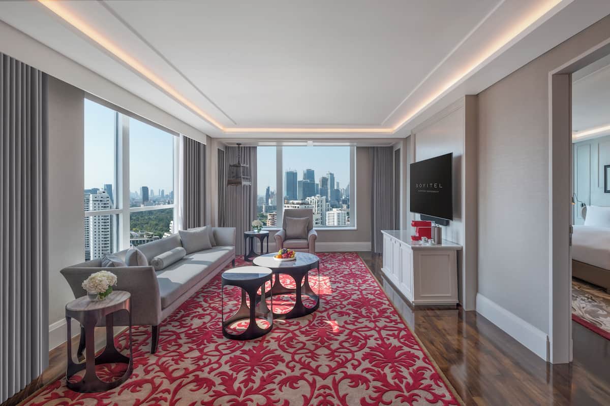A modern hotel room with a large window offering a city view, a sofa, a small table, and a TV.