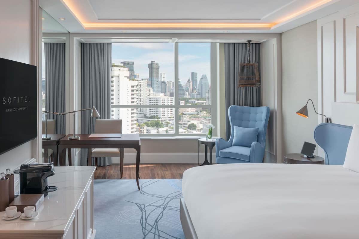 A hotel room with a large bed, a desk with a coffee maker, a chair, a TV, and a view of the city skyline.