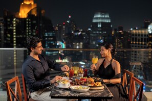 A couple dining at a table with a cityscape view, featuring a glass table, chairs, and a variety of dishes.