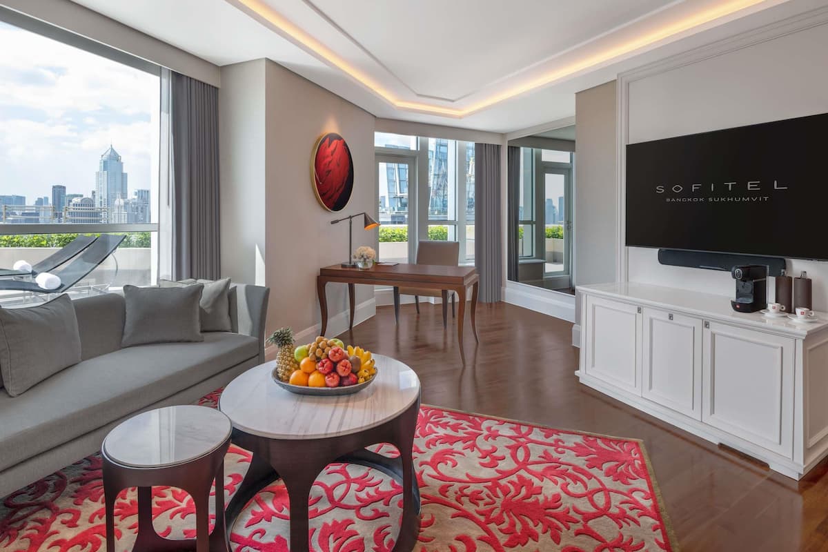 A modern hotel room with a sofa, dining table, fruit bowl, and a TV.