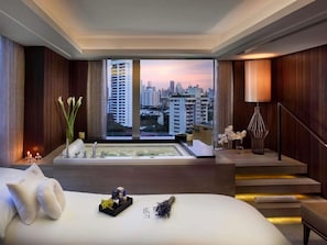A modern hotel room with a large bed, a bathtub, a view of the cityscape, and a staircase.