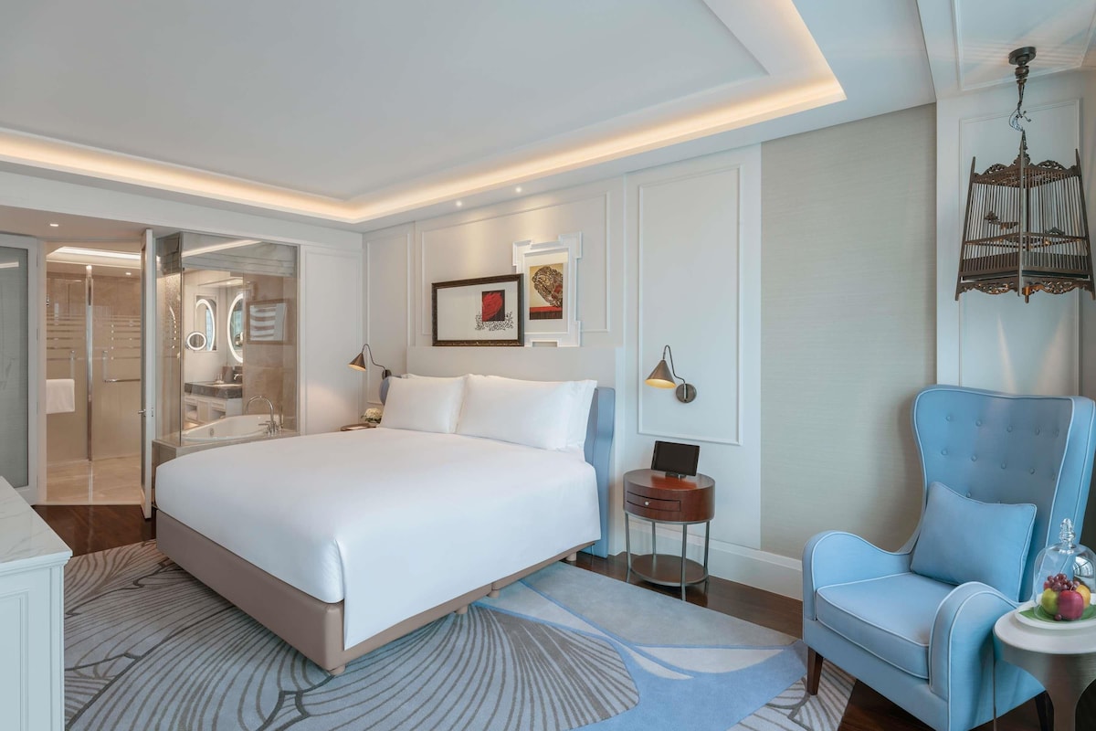 A modern hotel room with a large bed, a seating area with a blue chair, a small side table, and a shower area with glass doors.