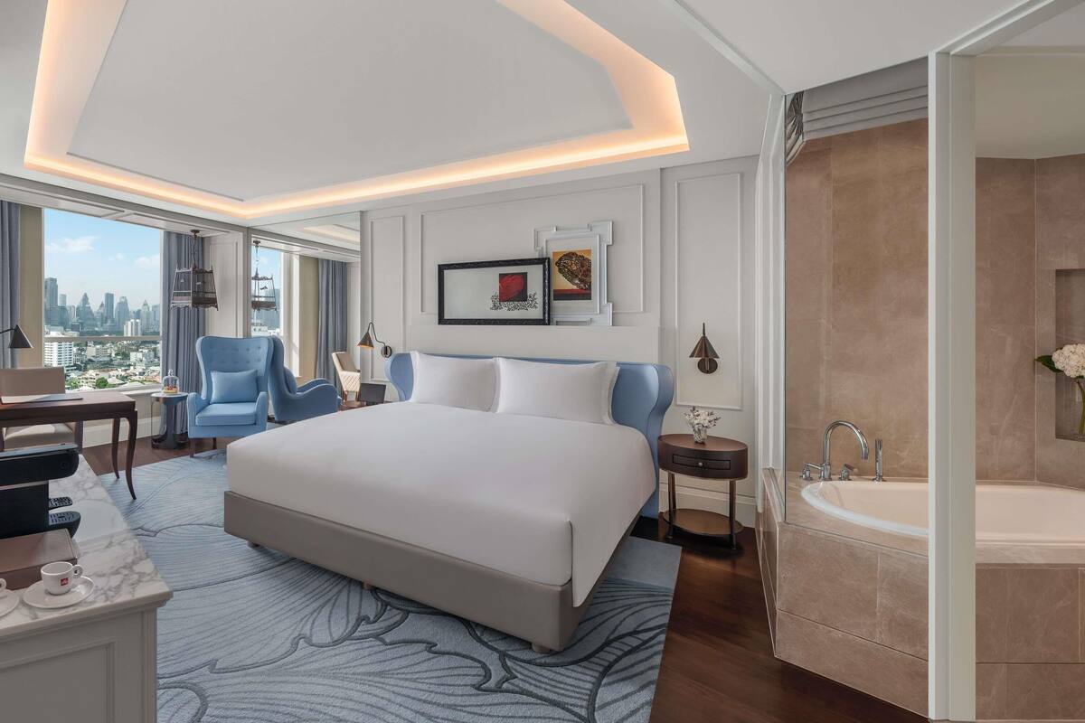 A modern hotel room with a large bed, a desk with a laptop, a chair, a view of the city, and a bathroom with a bathtub.
