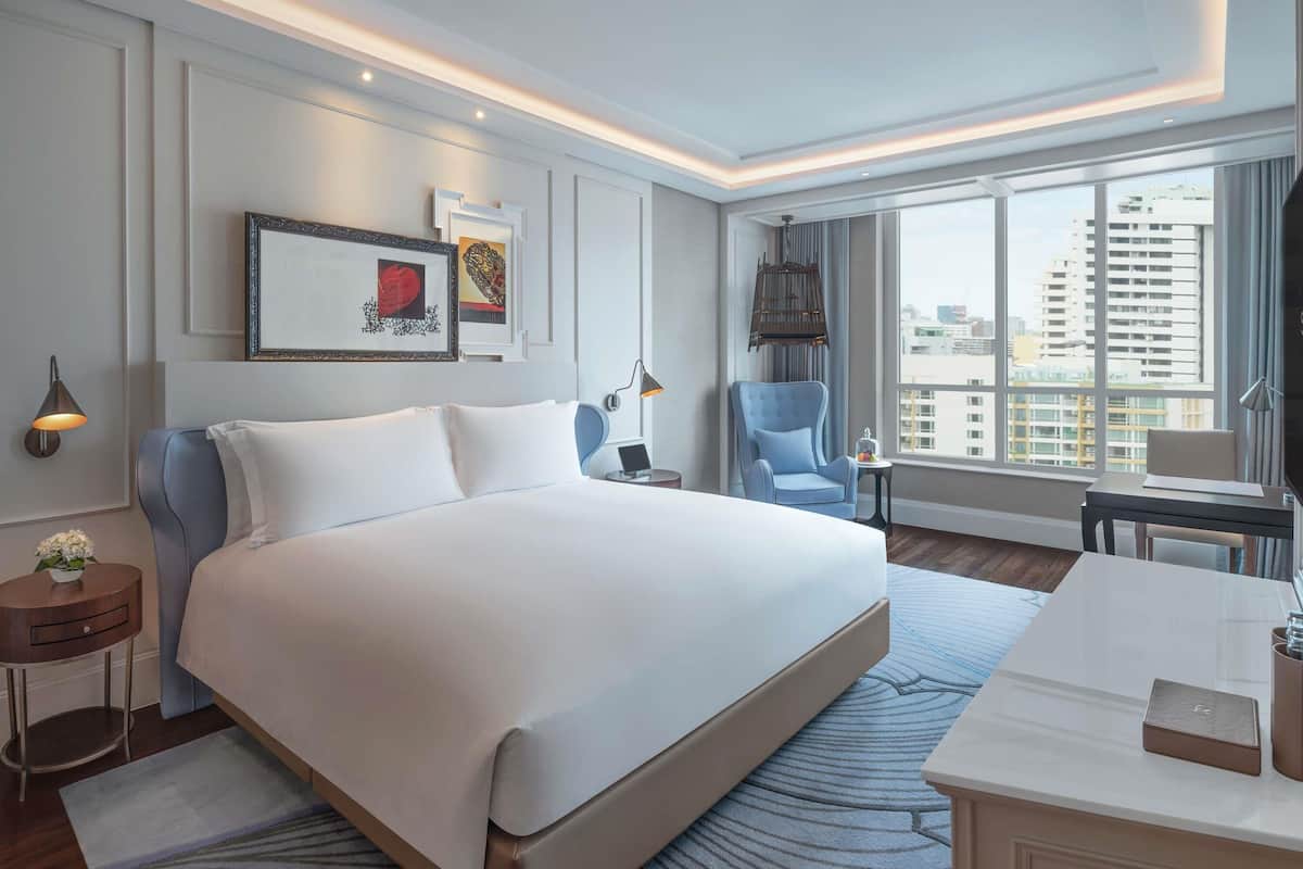 A hotel room with a large bed, a desk, a chair, a view of the city, and artwork on the wall.
