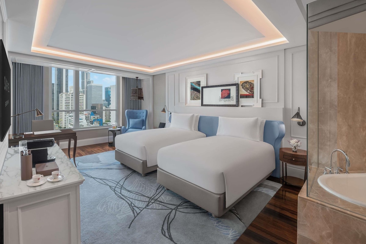A hotel room with two beds, a desk with a computer, a TV, a large window with a city view, and a bathroom with a bathtub.