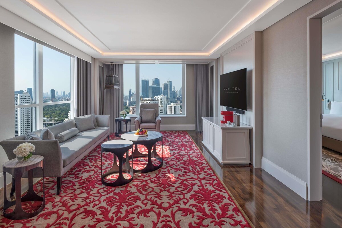 A modern hotel room with a large window, a sofa, two side tables, a TV, and a red patterned carpet.